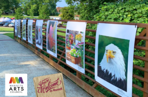 A display of artwork hangs along a rust-colored fence in an outdoor park setting, featuring various paintings including a bald eagle, flowers, and abstract designs. A Marietta Arts Council logo is visible.