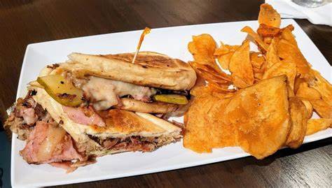 A sandwich with melted cheese, pickles, and meat is served on a plate with a side of potato chips.