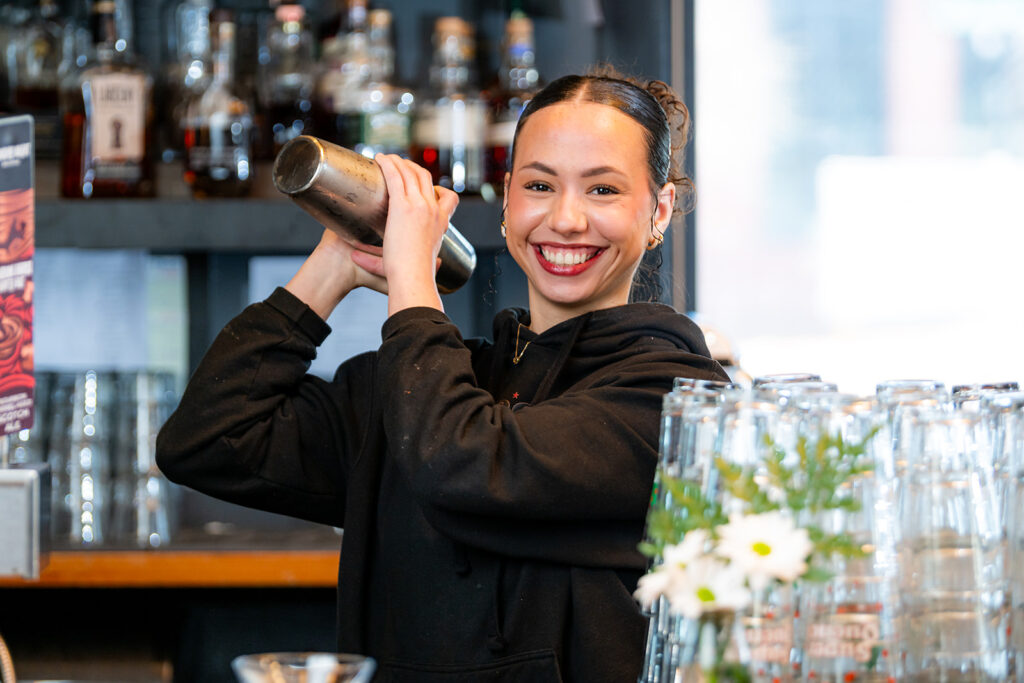 A smiling bartender wearing a black hoodie shakes a cocktail shaker behind a bar, with glassware and bottles visible in the background.