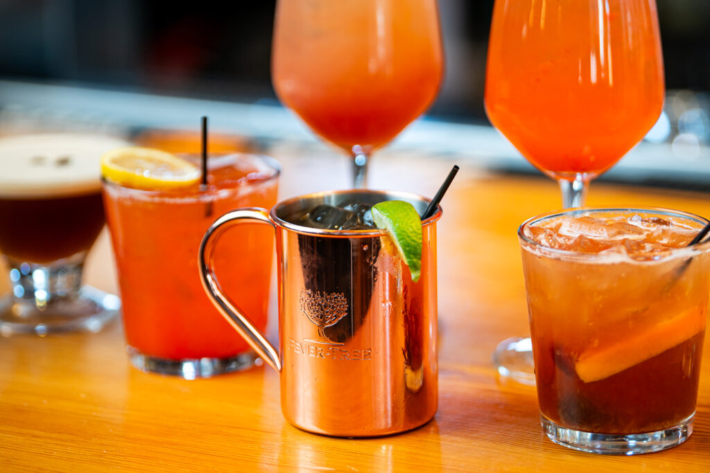 A variety of colorful cocktails in different glasses, including a copper mug with a lime wedge, are arranged on a wooden surface.