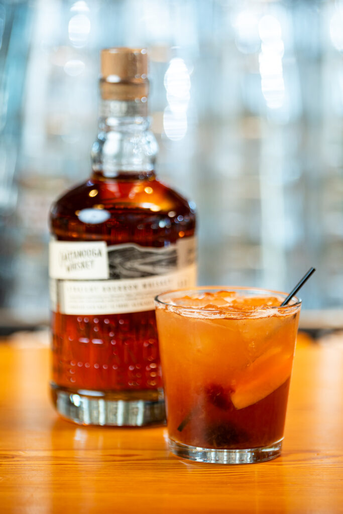 A bottle of Chattanooga Whiskey and a cocktail garnished with an orange slice and a cherry on a wooden surface.