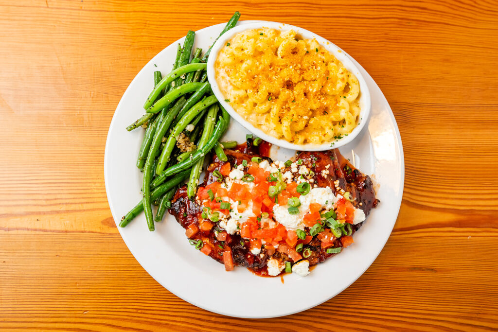 A white plate with green beans, macaroni and cheese, and a chicken breast topped with tomato, green onion, and cheese, on a wooden table.