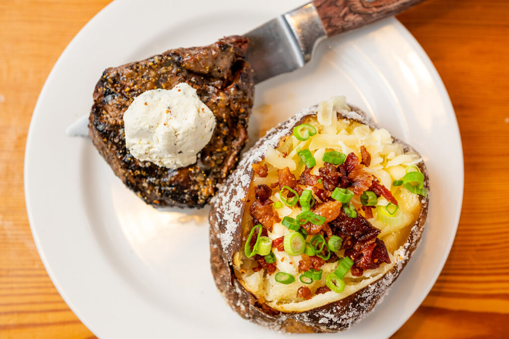 A grilled steak topped with a dollop of herb butter and a baked potato loaded with cheese, bacon, and green onions on a white plate.