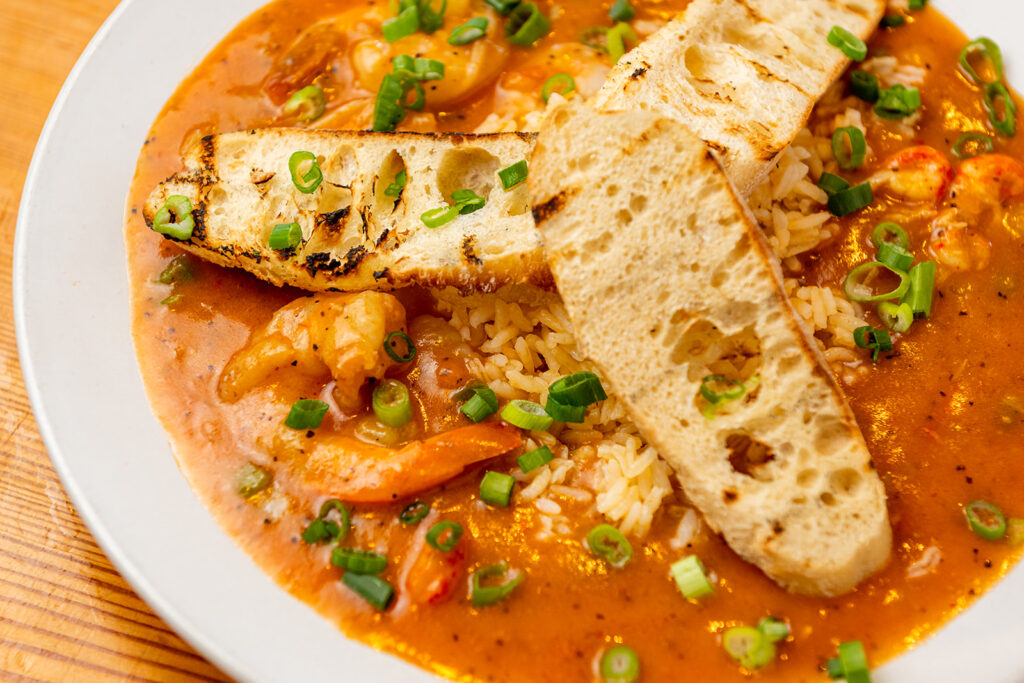 A plate of rice topped with shrimp and crawfish in a reddish sauce, garnished with chopped green onions and served with two pieces of grilled bread.