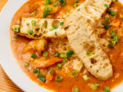 1885 Grill A plate of rice topped with shrimp and crawfish in a reddish sauce, garnished with chopped green onions and served with two pieces of grilled bread.