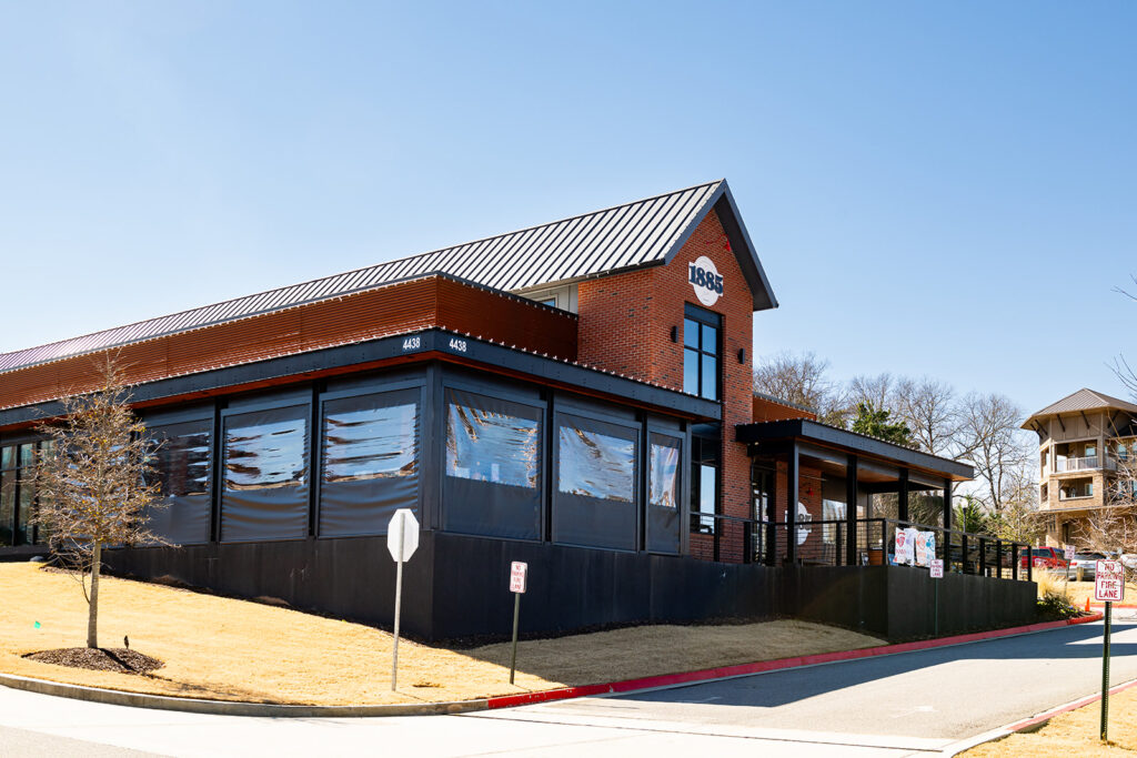 A modern brick restaurant building with large windows and a covered outdoor patio, located on a sloped lot with a sidewalk and street in front.