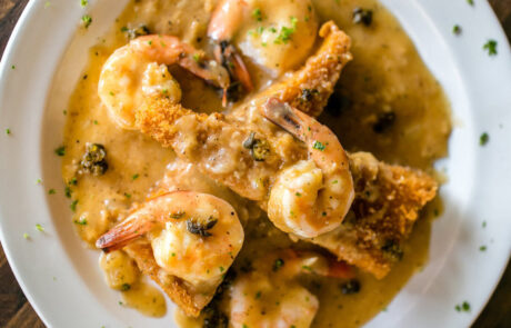 A white plate with shrimp, fried fish pieces, and a creamy brown sauce garnished with herbs.