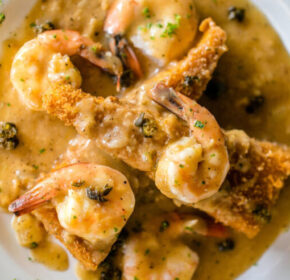 A white plate with shrimp, fried fish pieces, and a creamy brown sauce garnished with herbs.