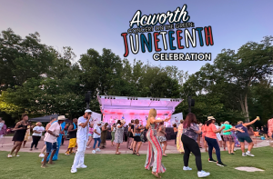 A crowd dances in front of a stage at an outdoor event, with trees in the background and a sign reading "Acworth Concert on the Green: Juneteenth Celebration" overhead.