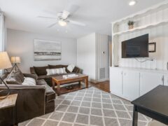 A living room with gray walls, a ceiling fan, and a geometric patterned rug. It features a brown couch, wooden coffee table, wall-mounted TV, and built-in shelving with decorative items.