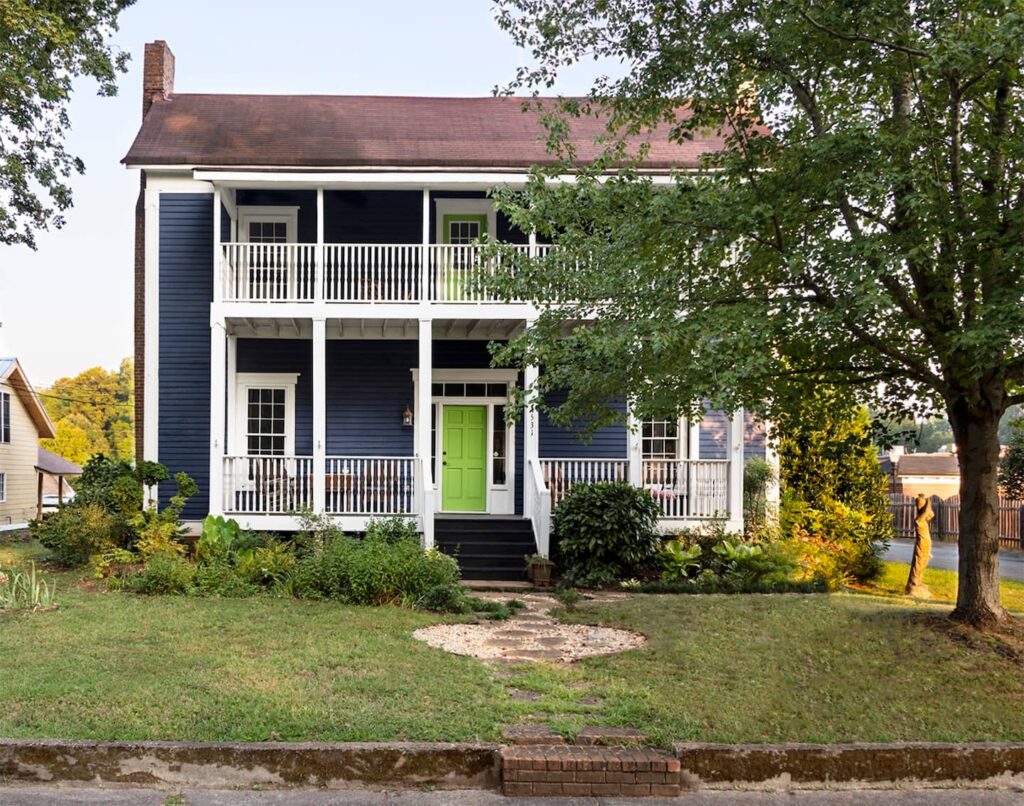 Two-story blue house with a green front door, white trim, and a double-decker porch. The home is surrounded by a grassy lawn and trees. Steps lead up to the front porch from the sidewalk.