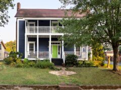 Two-story blue house with a green front door, white trim, and a double-decker porch. The home is surrounded by a grassy lawn and trees. Steps lead up to the front porch from the sidewalk.