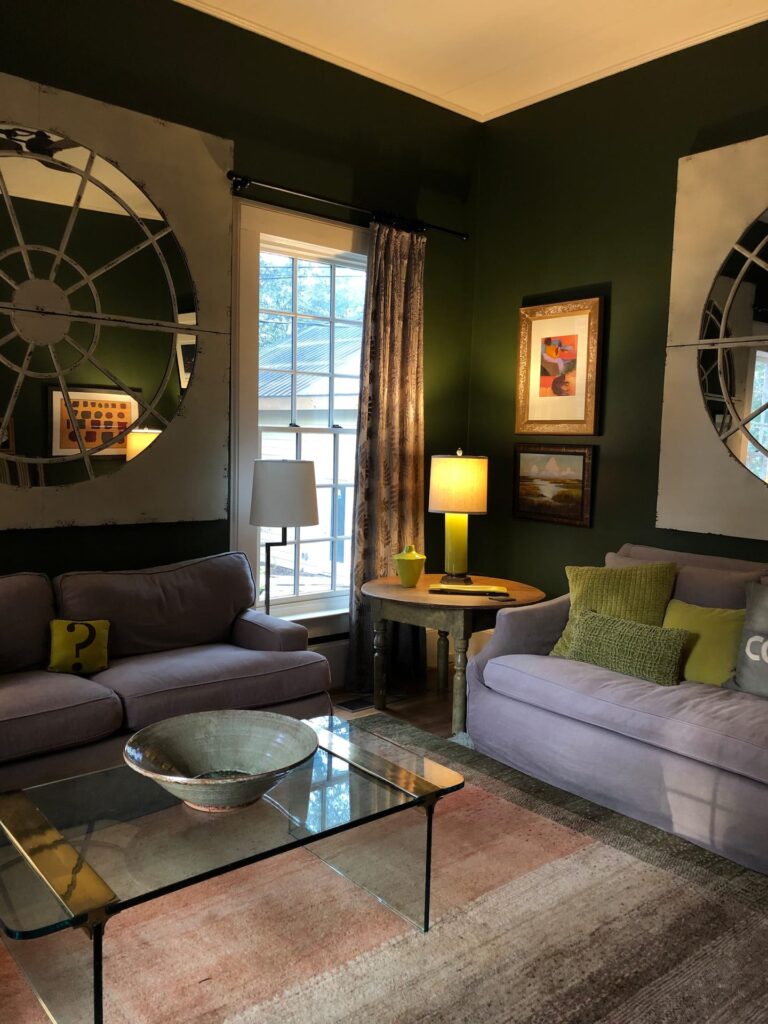 A living room with dark green walls, large mirrors, two grey couches, a glass coffee table, and various framed artworks. A window with drapes lets in natural light, illuminating the space.