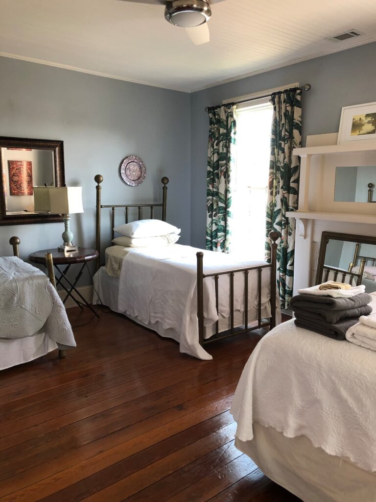 A bedroom with three single beds, a small table with a lamp, a mirror, and a window with leafy curtains. The walls are painted light gray and the floor is wooden.