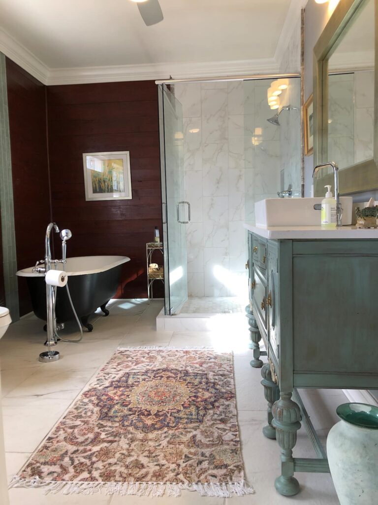 A bathroom with a freestanding black bathtub, glass-enclosed shower, large wooden vanity with a countertop sink, and an ornate rug on the white-tiled floor. A painting hangs on a wooden accent wall.