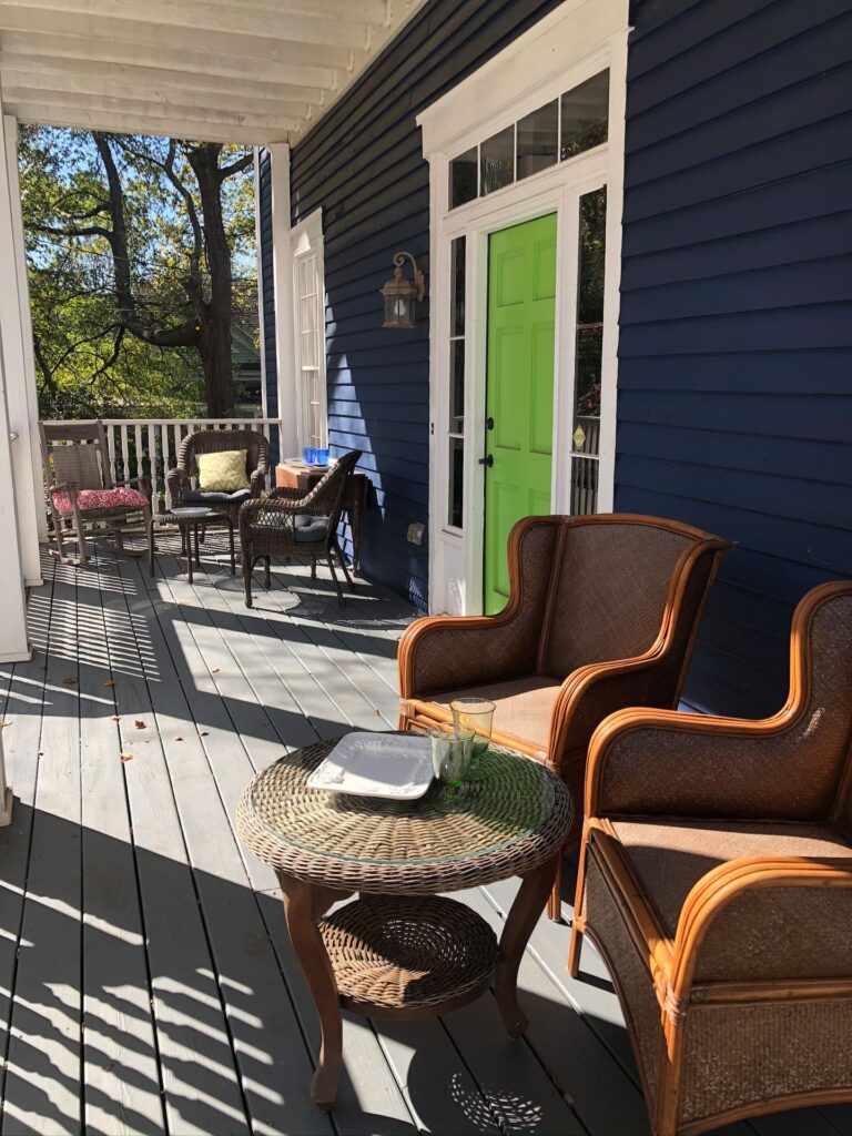 A porch with wooden flooring features wicker chairs, a round wicker table, and additional seating. A vibrant green door contrasts against the dark blue house exterior. Trees are visible in the background.