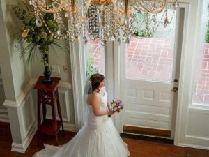 A bride in a white wedding gown stands indoors near a glass door, holding a bouquet and gazing outside. A chandelier hangs from the ceiling above.