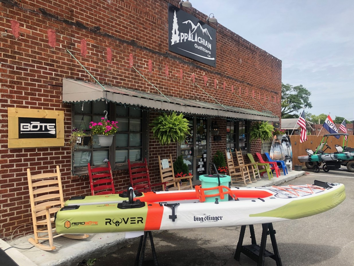 A storefront with a sign "Appalachian Outfitters." In front, a large kayak labeled "Rover," surrounded by chairs, and American flags displayed. Other outdoor gear is visible nearby.