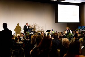 A man stands at a podium speaking to a crowded room during an event. Large screens in the background display information. The audience sits at tables decorated with floral arrangements.