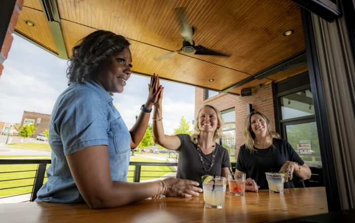 Three women sit at a table with drinks, two sharing a high five, enjoying an outdoor setting with city views—celebrating the best of Cobb County with their Cobb Foodie Week Meal Deals Pass!.