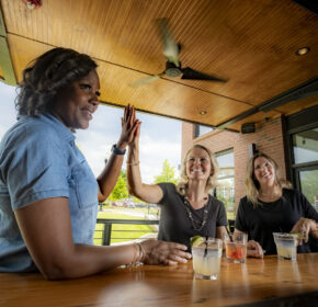Three women sit at a table with drinks, two sharing a high five, enjoying an outdoor setting with city views—celebrating the best of Cobb County with their Cobb Foodie Week Meal Deals Pass!.
