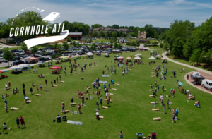 Aerial view of a Cornhole ATL tournament on a large grassy field with numerous games in progress and a crowded parking lot in the background.