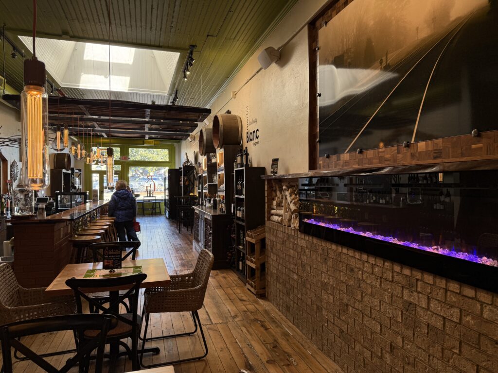 Cozy cafe interior with wooden floor, tables, and chairs. A person stands near the counter. A large photo and electric fireplace adorn the wall. Natural light streams through skylights.