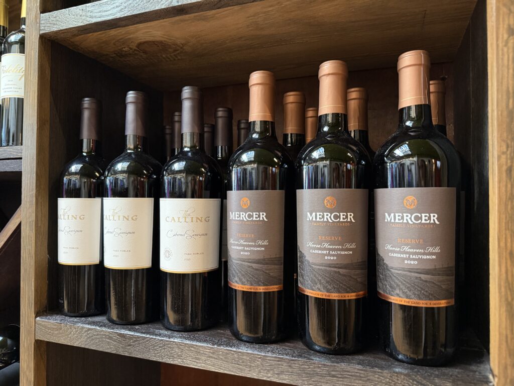 Bottles of red wine, including The Calling and Mercer labels, displayed on a wooden shelf.