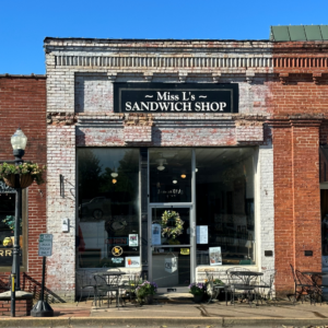 A brick storefront with a sign reading "Miss L's Sandwich Shop" beckons hungry hikers from the list of Six Great Places to Eat in Acworth After A Hike. Outdoor seating features metal tables and chairs, while a potted plant hangs invitingly by the entrance. A lamppost stands sentinel on the left.