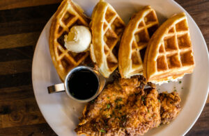 A plate with four waffle quarters, a dollop of butter, a small cup of syrup, and pieces of fried chicken on a wooden table; foodies and fun-seekers flock to Acworth for this mouthwatering delight.