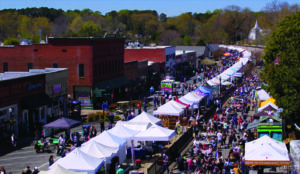 Aerial view of a bustling street market with numerous white tents and large crowds of people; brick buildings line one side of the street, and trees and a white church spire are visible in the background—just one of Five Reasons Acworth is Atlanta's Go-To Festival Spot.