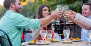 Three people sitting at an outdoor dining table clink glasses while smiling. Plates of food and water glasses are on the table. Trees and a railing are visible in the background, reflecting why FOODIES AND FUN-SEEKERS FLOCK TO ACWORTH for such delightful gatherings.