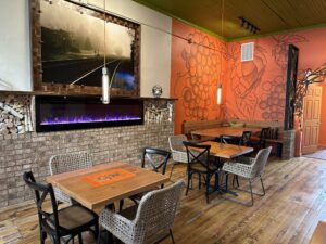 A cozy cafe interior with wooden tables and chairs, a brick wall with a lit electric fireplace below a large framed photo, and a mural of grapes on an orange wall awaits foodies and fun-seekers flocking to Acworth.