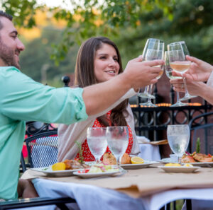 Three people sitting at an outdoor restaurant in Acworth clink wine glasses in a toast. Plates of food and water glasses are on the table, adding to the festive atmosphere. Trees are visible in the background as foodies and fun-seekers flock to this delightful spot.