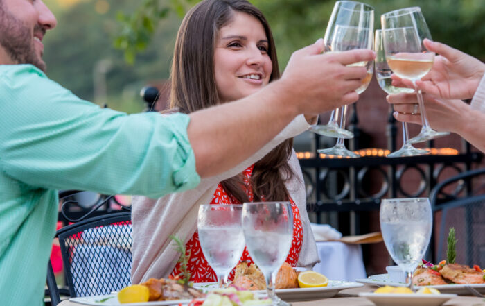 Three people sitting at an outdoor restaurant in Acworth clink wine glasses in a toast. Plates of food and water glasses are on the table, adding to the festive atmosphere. Trees are visible in the background as foodies and fun-seekers flock to this delightful spot.