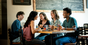 Five young adults sit around a table at a restaurant, laughing and eating. Menus are visible on the wall behind them as they appear to be enjoying pizza and canned drinks—just the kind of lively scene that shows why foodies and fun-seekers flock to Acworth.