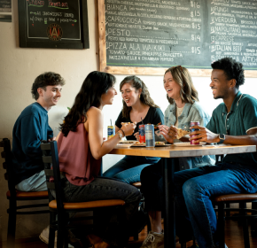Five young adults sit around a table at a restaurant, eating and drinking while talking and smiling. Chalkboard menus with food items are visible on the wall behind them—a lively scene that shows why foodies and fun-seekers flock to Acworth.