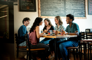 Five young adults sit around a table at a restaurant, eating and drinking while talking and smiling. Chalkboard menus with food items are visible on the wall behind them—a lively scene that shows why foodies and fun-seekers flock to Acworth.