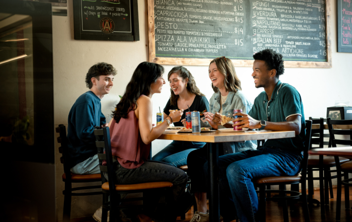 Five young adults sit around a table at a restaurant, eating and drinking while talking and smiling. Chalkboard menus with food items are visible on the wall behind them—a lively scene that shows why foodies and fun-seekers flock to Acworth.
