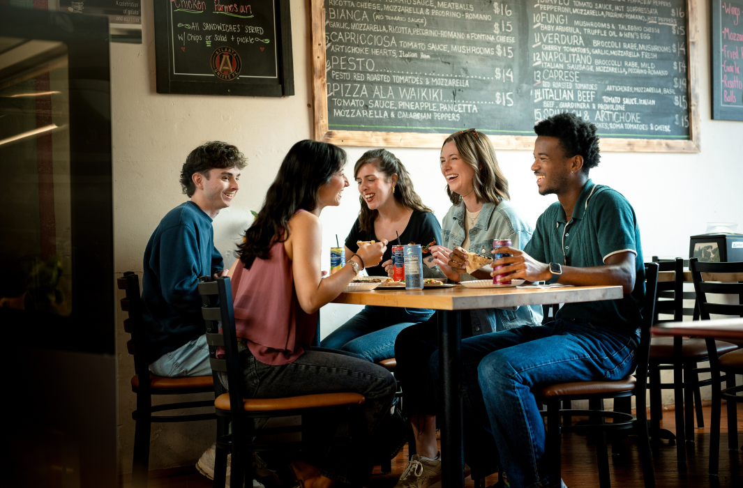 Five young adults sit around a table at a restaurant, eating and drinking while talking and smiling. Chalkboard menus with food items are visible on the wall behind them—a lively scene that shows why foodies and fun-seekers flock to Acworth.