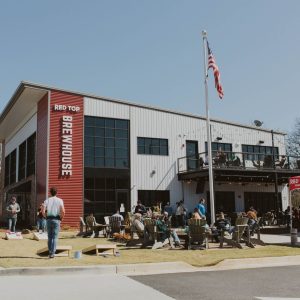 People sit and stand outside a two-story brewhouse with a large sign reading "Red Top Brewhouse," playing lawn games and socializing under sunny skies. An American flag is raised nearby—Celebrate Your Event in Acworth!.