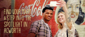 Two people take a selfie in front of a mural with text that reads, "Find Your Mark & Step Into the Spotlight in Acworth.