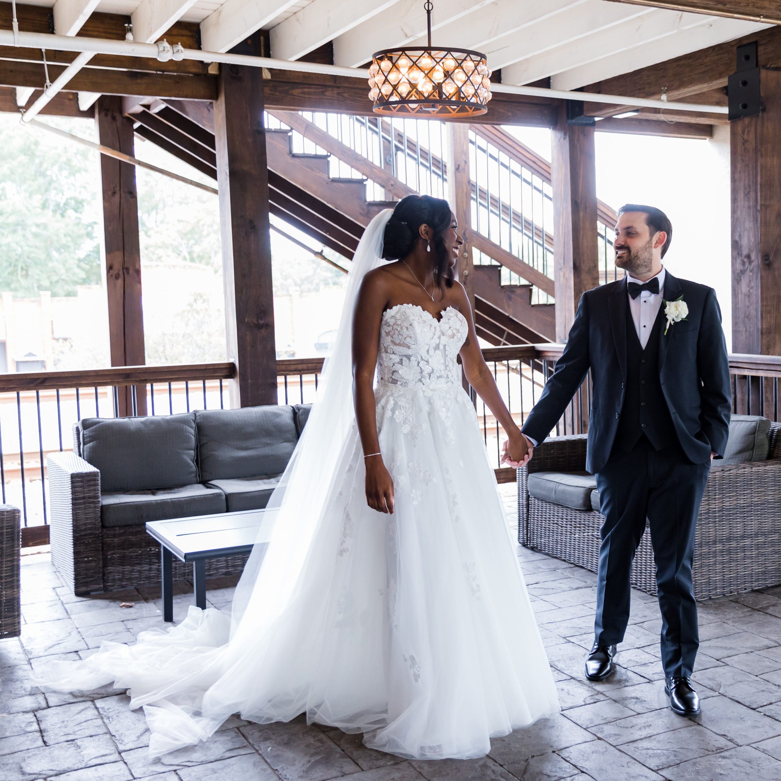 A bride in a white gown and a groom in a black tuxedo stand indoors on a patio, holding hands and looking at each other—Celebrate Your Event in Acworth!.