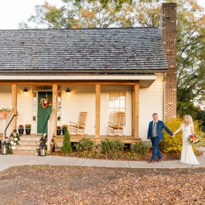 A bride and groom hold hands while walking in front of a rustic house with a porch, rocking chairs, and autumn decorations—Celebrate Your Event in Acworth!.