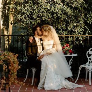 A bride in a white gown and a groom in a dark suit share a kiss while sitting on a white chair on a brick patio with greenery and red roses behind them. Celebrate Your Event in Acworth for unforgettable moments like this!.