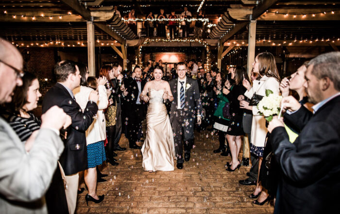 A bride and groom walk through a crowd of cheering guests under string lights, with bubbles floating around, as foodies and fun-seekers flock to Acworth for the celebration.