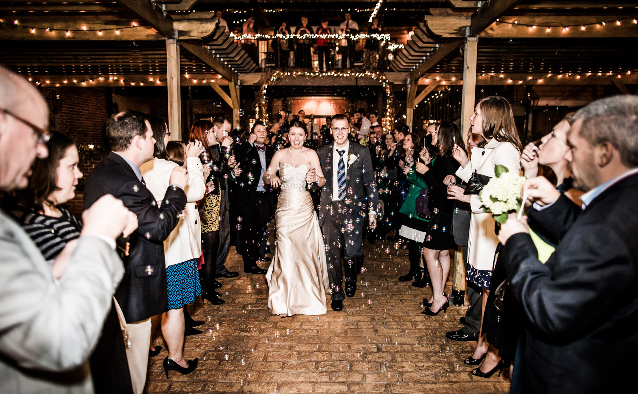 A bride and groom walk through a crowd of cheering guests under string lights, with bubbles floating around, as foodies and fun-seekers flock to Acworth for the celebration.