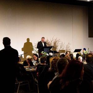 A man stands at a podium addressing a seated audience during an indoor formal event, with floral arrangements and table settings—Celebrate Your Event in Acworth!.