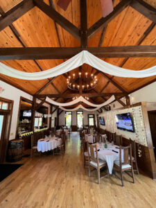 A rustic dining room with a wooden ceiling and chandeliers, where foodies and fun-seekers flock to Acworth. Tables are set with white tablecloths, surrounded by wooden chairs, and draped fabric decorates the interior.