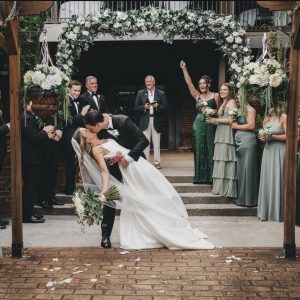 A bride and groom kiss under a floral arch while wedding party members in green and gray stand and celebrate around them. Celebrate Your Event in Acworth!.